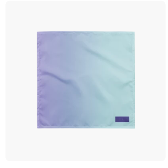 A square fabric pocket square or scarf with a smooth gradient transitioning from lavender purple on the left to light aqua blue on the right, featuring a small dark purple rectangular brand tab in the bottom right corner.