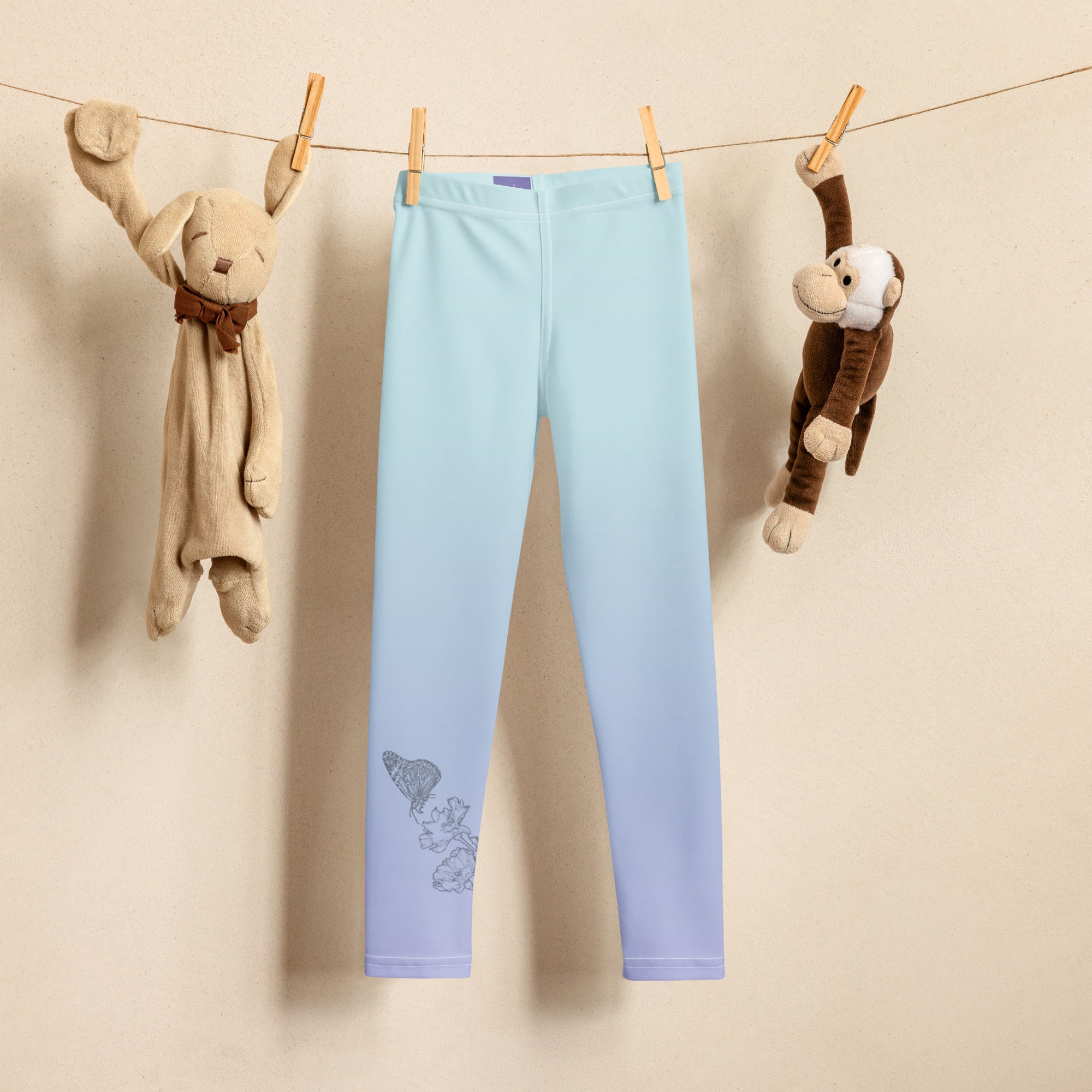 Light blue gradient children's leggings hanging from a clothesline with wooden clothespins, flanked by plush toys against a beige wall.