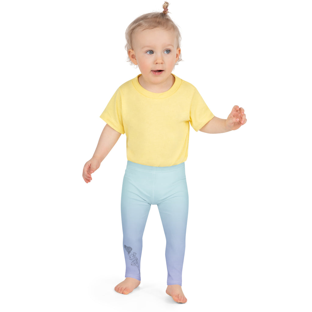 A toddler standing against a white background, wearing a yellow short-sleeve t-shirt and light blue leggings with a subtle dark graphic on one leg.