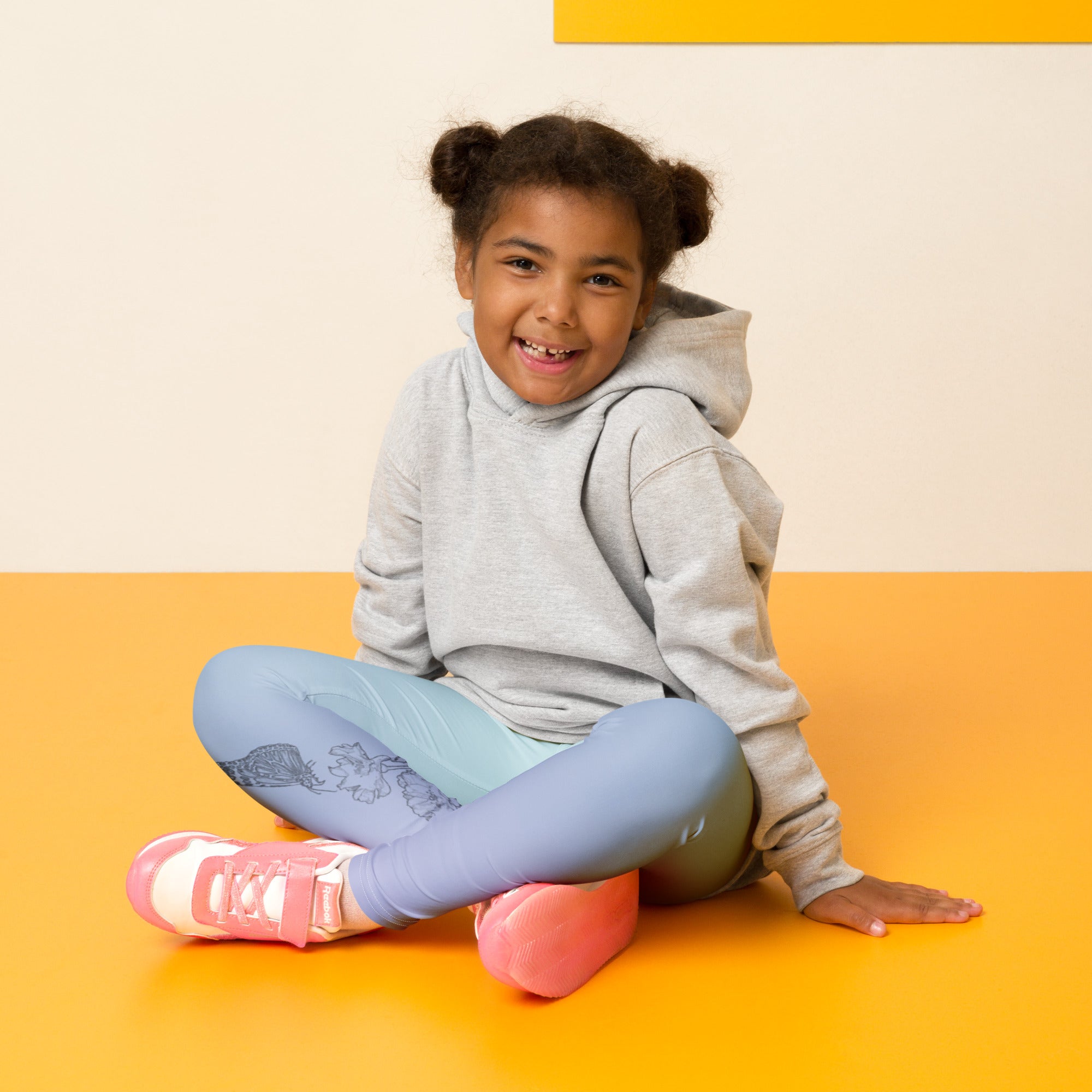 A young girl with her hair in two buns sits cross-legged on a bright yellow floor against a cream wall. She is wearing a light gray hoodie, light blue leggings with a dark graphic print, and pink sneakers.
