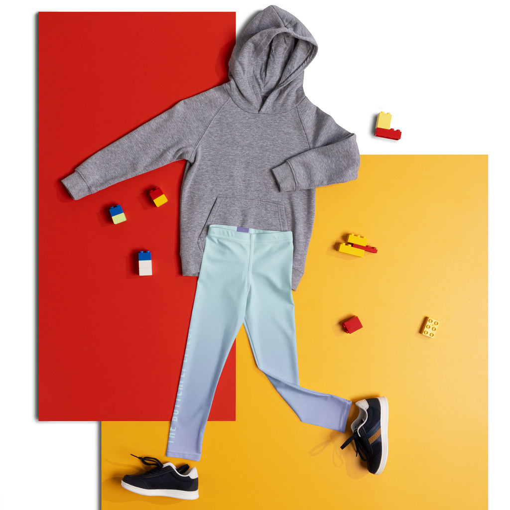 A child's clothing outfit laid flat on a split red and yellow background, featuring a heather gray hoodie, light blue leggings, and black sneakers, with small toy blocks scattered around.