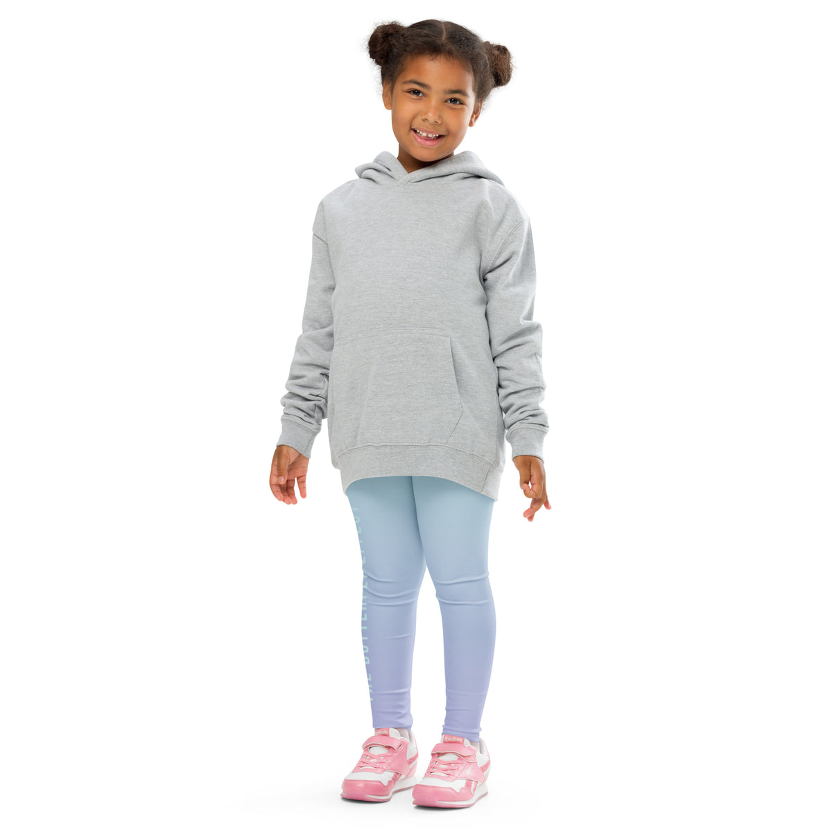 A young girl with her hair in two buns stands against a white background wearing a light gray oversized pullover hoodie, light blue leggings, and pink sneakers.
