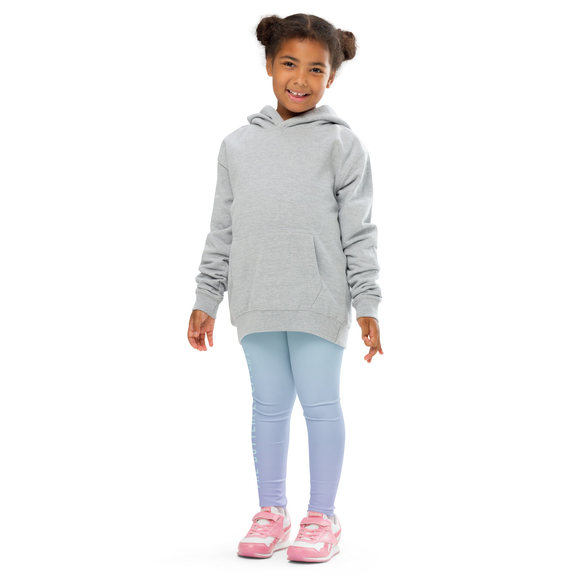 A young girl with her hair in two buns stands against a white background wearing a light gray oversized pullover hoodie, light blue leggings, and pink sneakers.