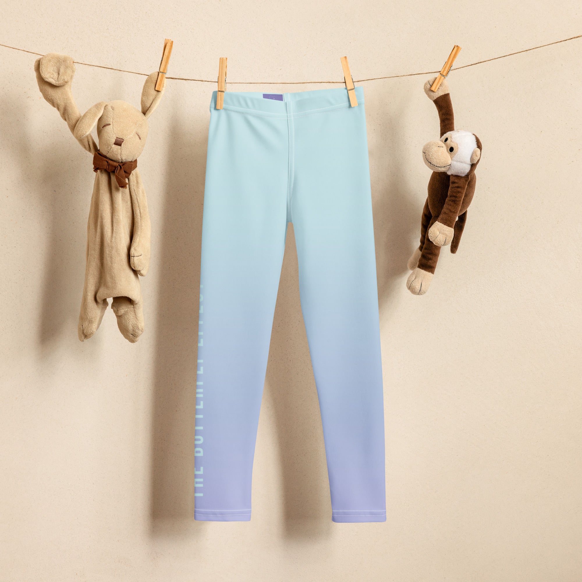 Light blue gradient leggings hanging from a clothesline with wooden clothespins, flanked by plush toys against a beige wall.