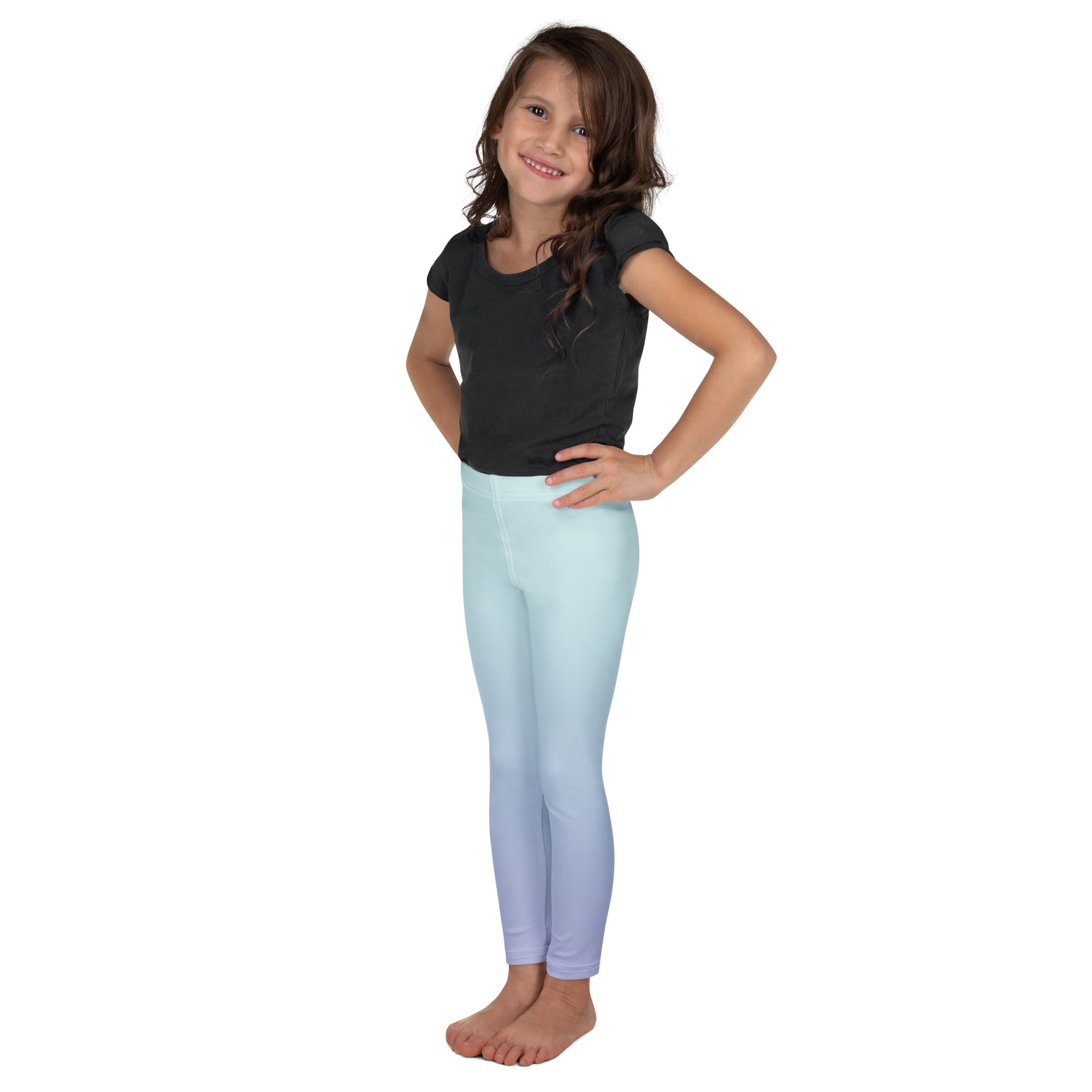 A young girl with long brown hair stands barefoot against a solid white background, wearing light blue gradient leggings with a subtle graphic print near the lower left leg and a dark gray short-sleeve t-shirt.