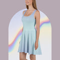 A woman wearing a light blue scoop-neck skater dress, standing in front of a white arched backdrop with a soft rainbow gradient.