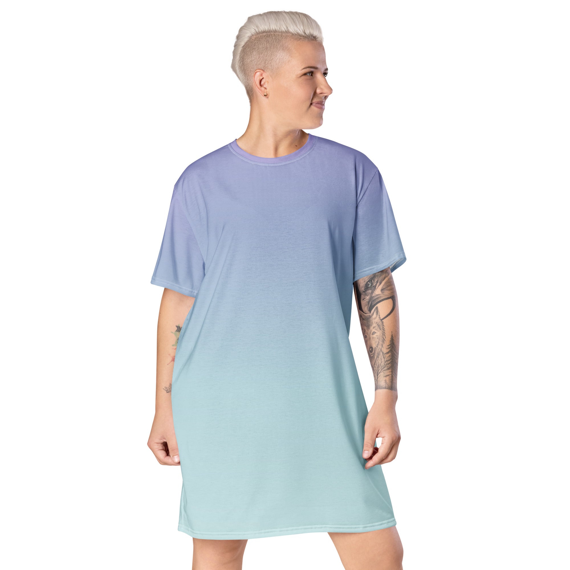 A model with short blonde hair and tattoos is shown from the back wearing a short-sleeve crewneck t-shirt dress with a smooth vertical gradient transitioning from lavender purple at the top to light mint green at the bottom, set against a plain white background.
