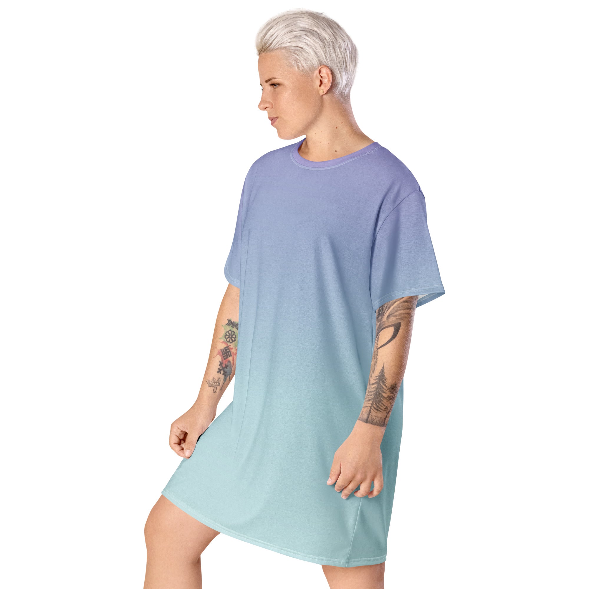 A model with short platinum blonde hair and tattoos is shown from the side wearing an oversized short-sleeve t-shirt dress with a smooth color gradient transitioning from periwinkle blue to light mint green, set against a plain white background.