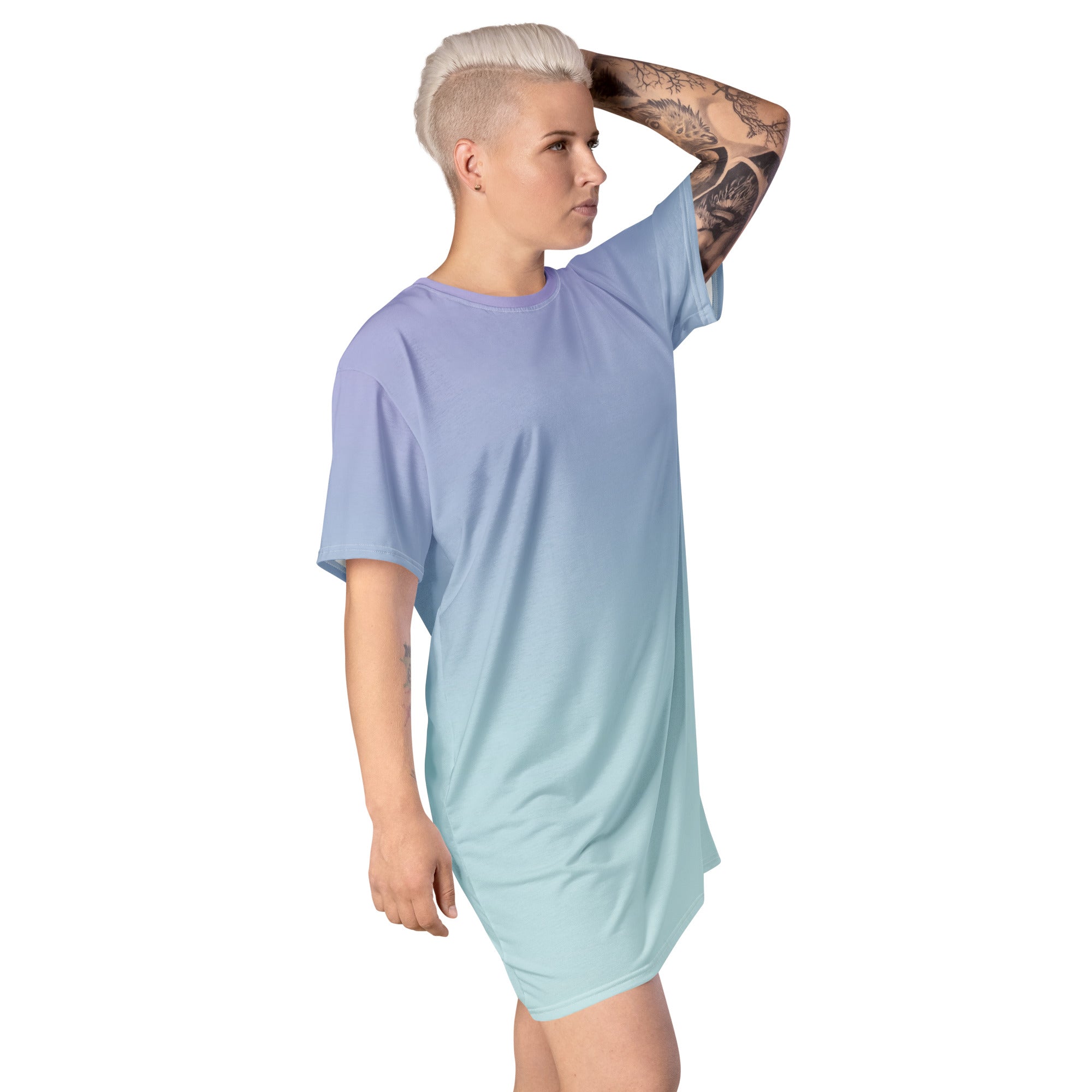 A model with short platinum blonde hair and a tattooed arm wearing a short-sleeve t-shirt dress with a smooth color gradient transitioning from lavender-purple at the top to light aqua-blue at the bottom, set against a plain white background.