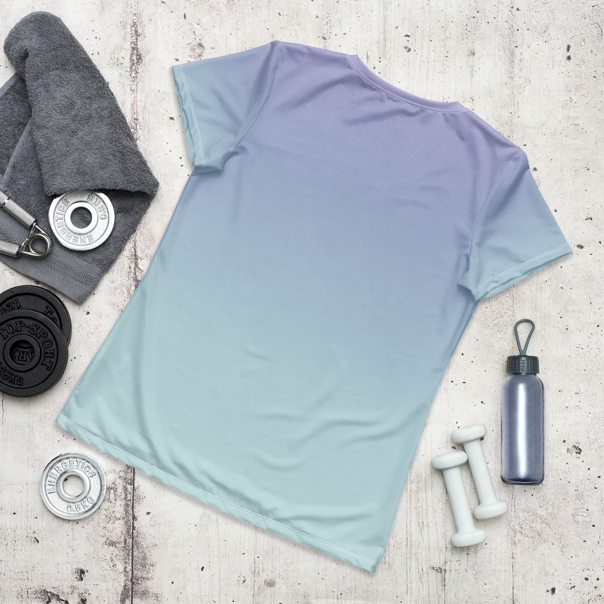 A short-sleeve athletic T-shirt laid flat on a light wood surface, featuring a smooth color gradient from lavender purple at the shoulders to a pale mint green at the hem. Styled with fitness props including a towel, dumbbells, a water bottle, and a barbell.