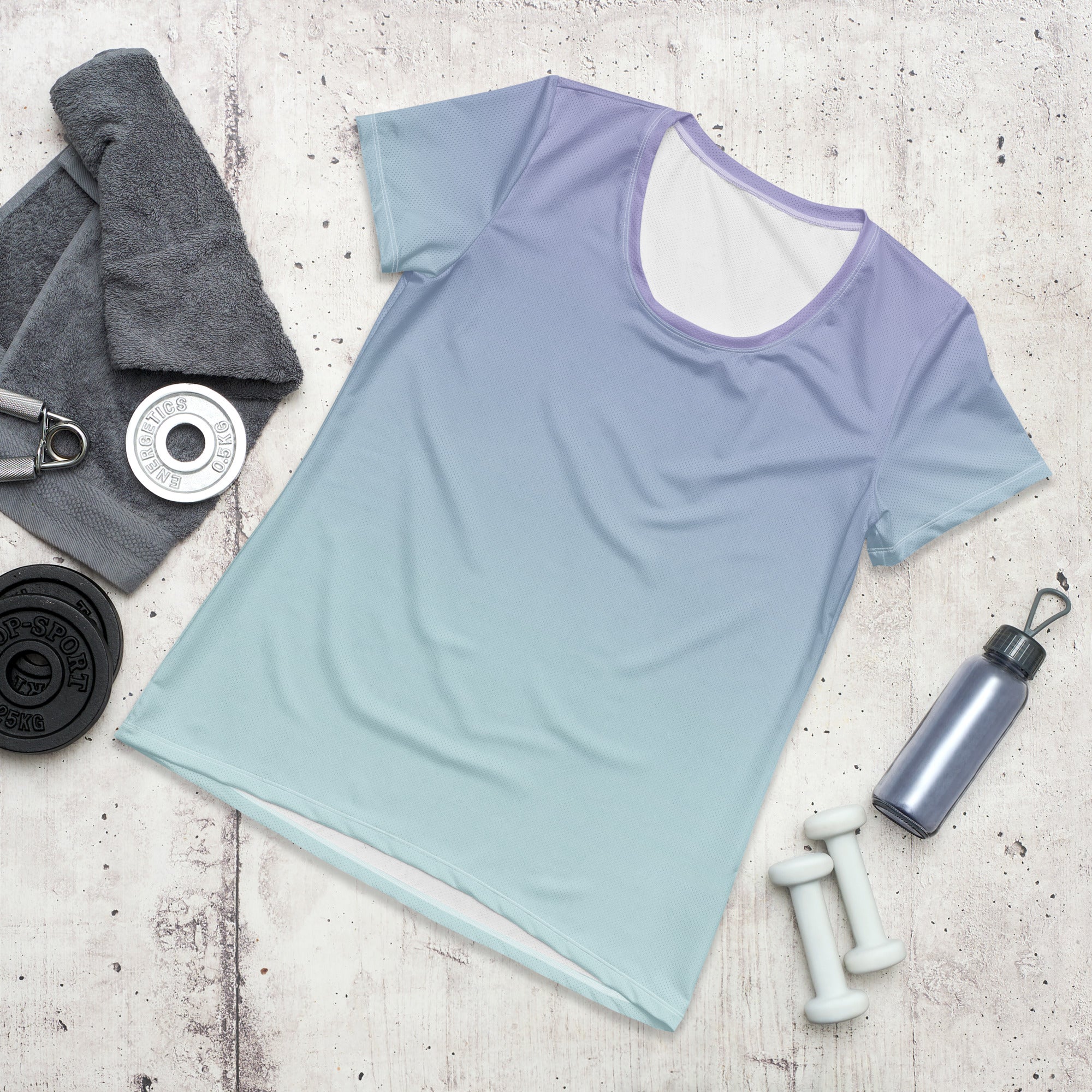 A short-sleeve athletic top with a smooth color gradient transitioning from lavender purple at the shoulders to a light mint green at the hem, styled in a flat lay with fitness props on a light concrete background.