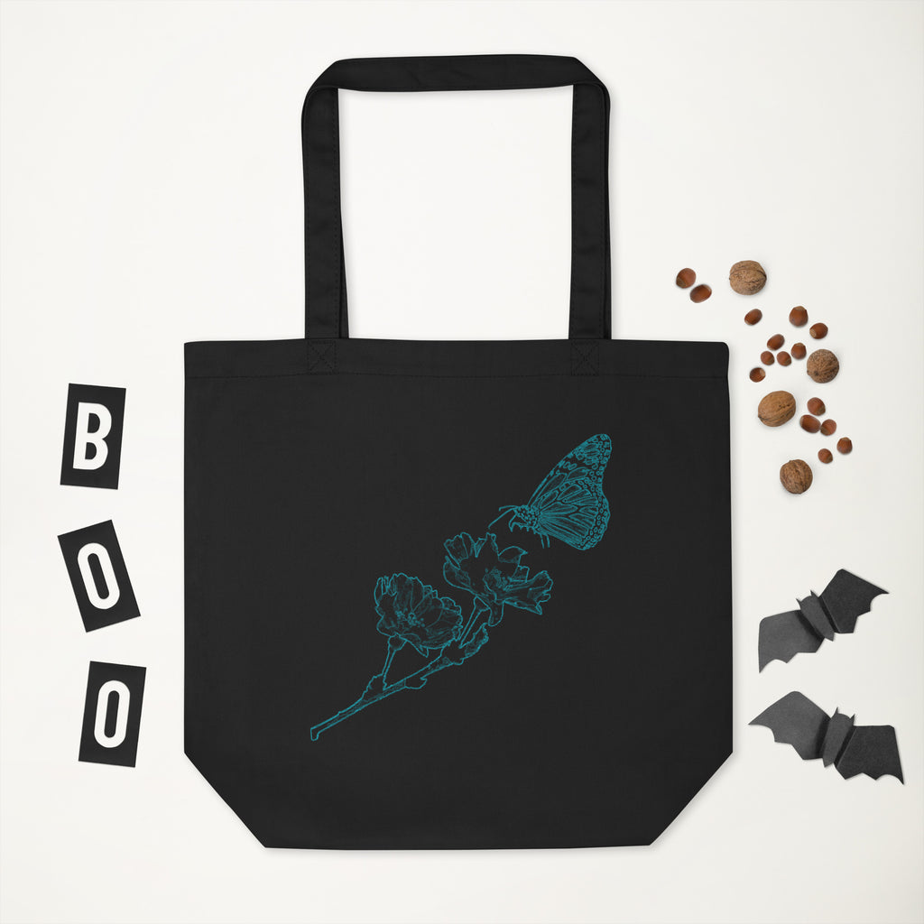 A black canvas tote bag with a teal butterfly and flower line art design, displayed on a white background with Halloween-themed props including 'BOO' letter blocks, cat food, and bat cutouts.