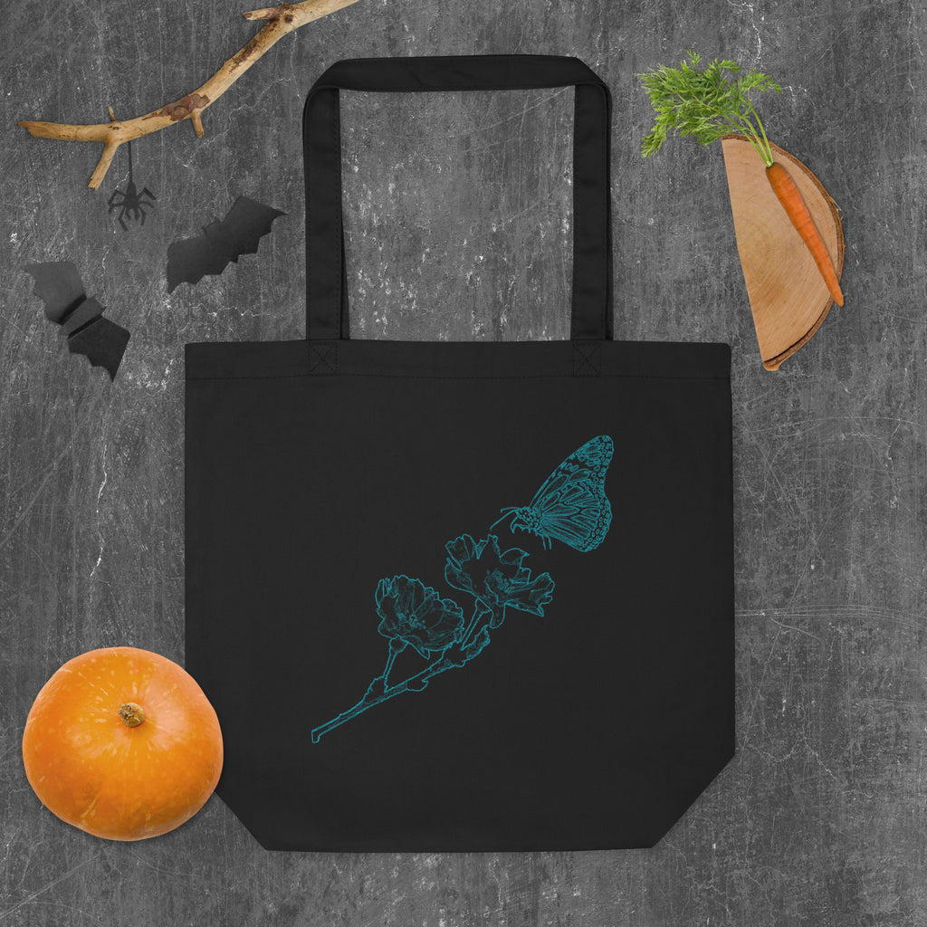 A black canvas tote bag with a teal butterfly and flower line-art design, styled flat-lay on a gray textured background with Halloween-themed props including a pumpkin, carrot, bat silhouette, and spider decoration.