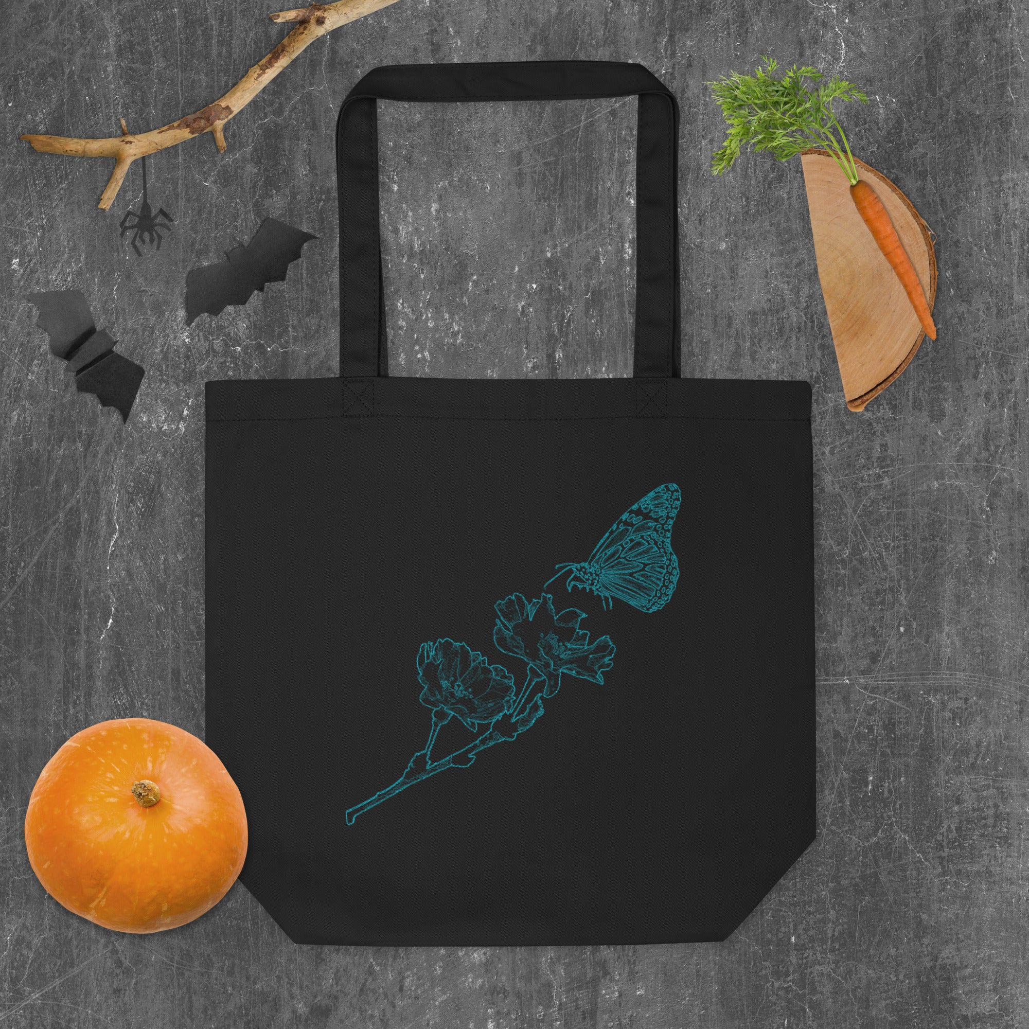 A black canvas tote bag with a teal butterfly and flower line-art design, styled flat-lay on a gray textured background with Halloween-themed props including a pumpkin, carrot, bat silhouette, and spider decoration.