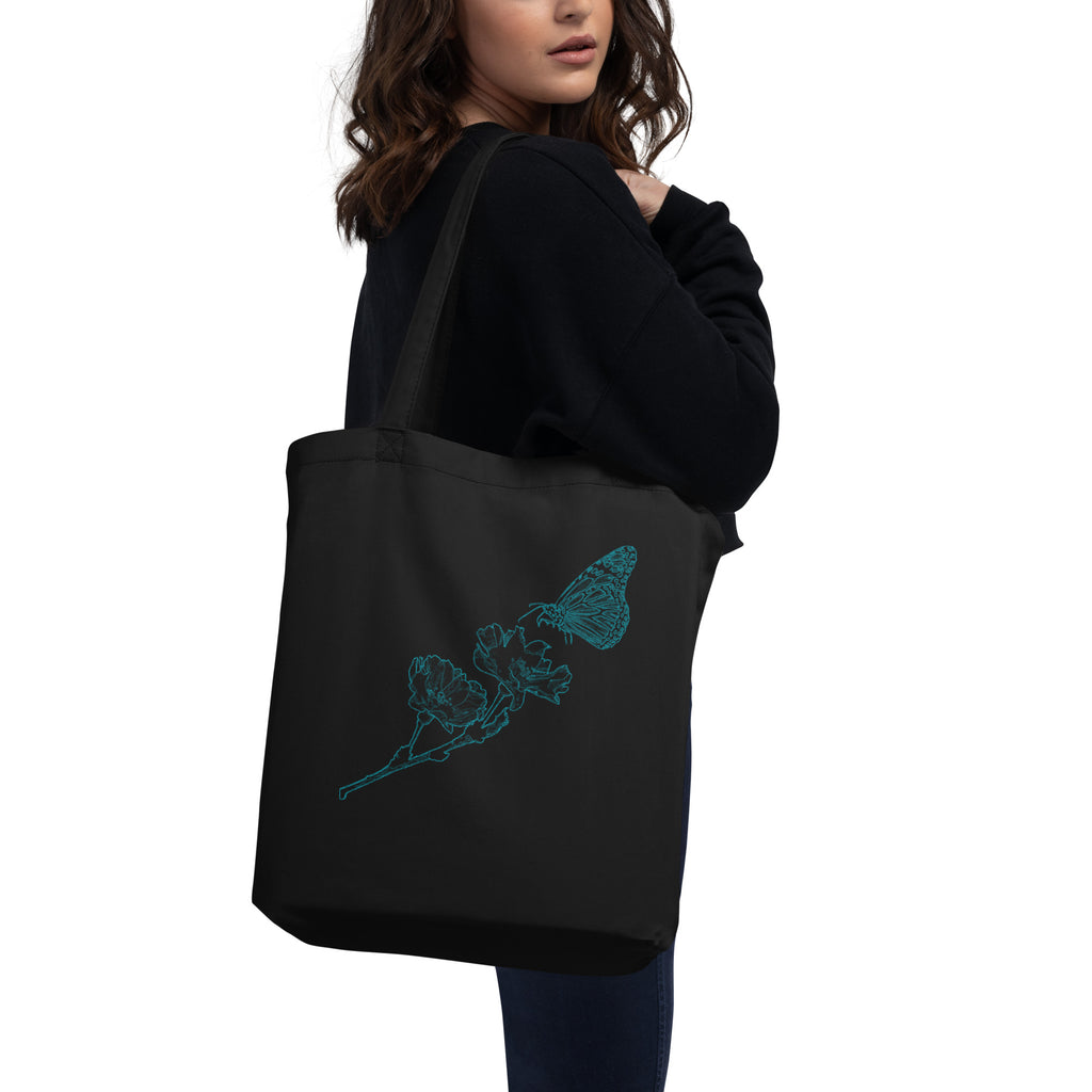 A woman wearing a black hoodie and blue jeans carries a black canvas tote bag with a white cartoon character drawing and a speech bubble on the front, against a plain white background.