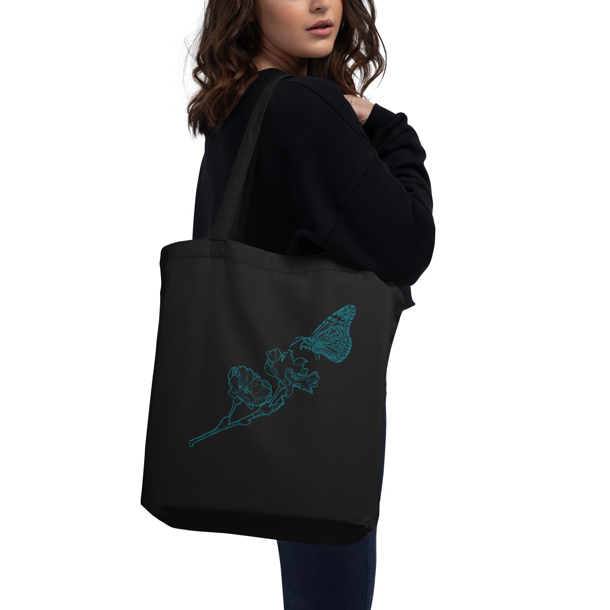 A woman wearing a black hoodie and blue jeans carries a black canvas tote bag with a white cartoon character drawing and a speech bubble on the front, against a plain white background.