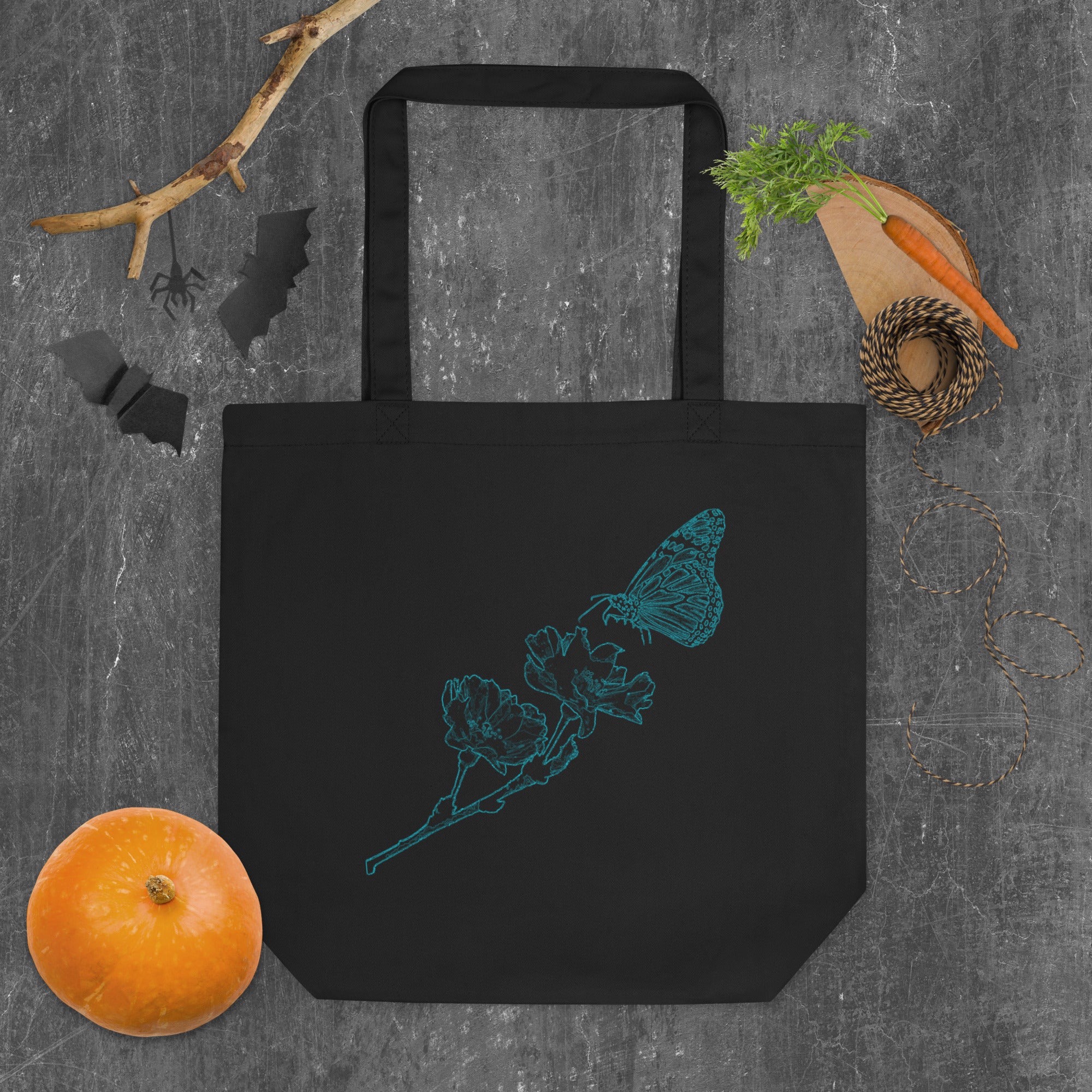 A black canvas tote bag with a teal butterfly and flower line-art design, styled in a flat-lay on a gray textured background with autumn props including a small orange pumpkin, a branch, black bat silhouettes, and twine.