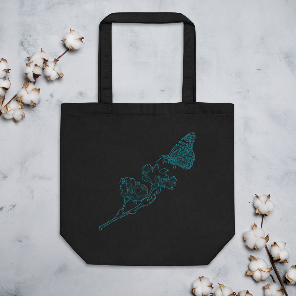 A black canvas tote bag with a teal butterfly and flower line-art design, displayed flat on a light gray marble surface with cotton bolls around it.