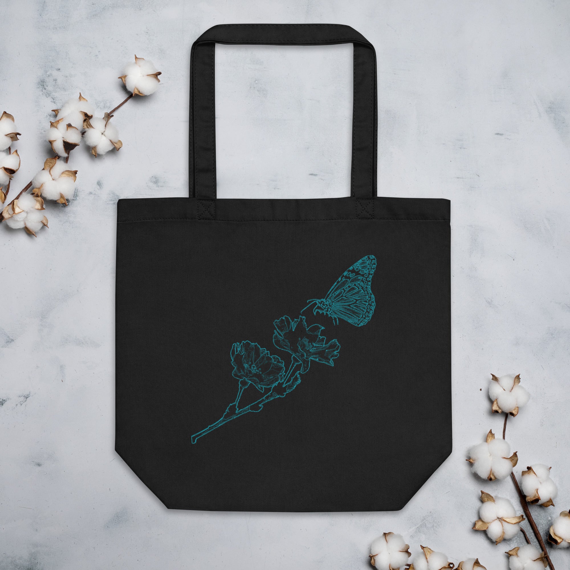 A black canvas tote bag with a teal butterfly and flower line-art design, displayed flat on a light gray marble surface with cotton bolls around it.