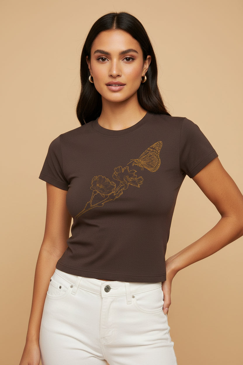 A woman modeling a dark brown short-sleeve crop top t-shirt with a gold butterfly and floral graphic on the front, paired with white jeans, set against a solid beige background.