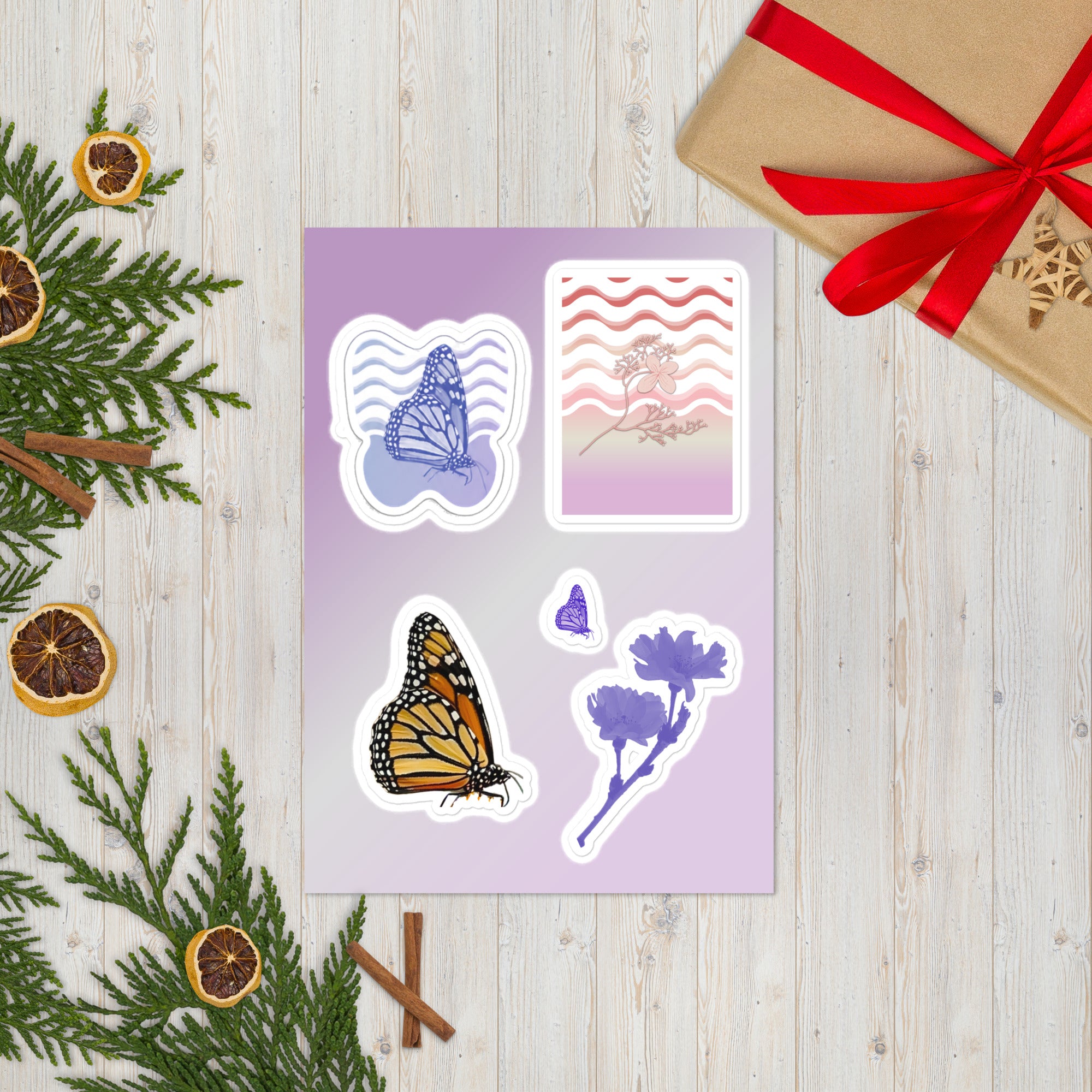 A sheet of four decorative stickers with butterfly, floral, and wavy line designs, styled on a light wood surface with greenery, dried orange slices, cinnamon sticks, and a wrapped gift box.