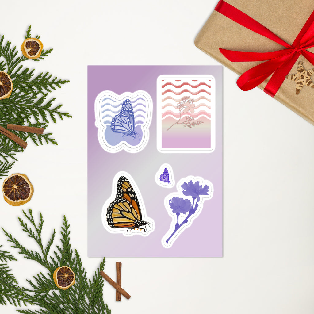 A sheet of decorative stickers displayed on a light background with holiday greenery, dried orange slices, cinnamon sticks, and a wrapped gift box. The stickers include a butterfly, a monarch butterfly, floral illustrations, and a patterned card design.