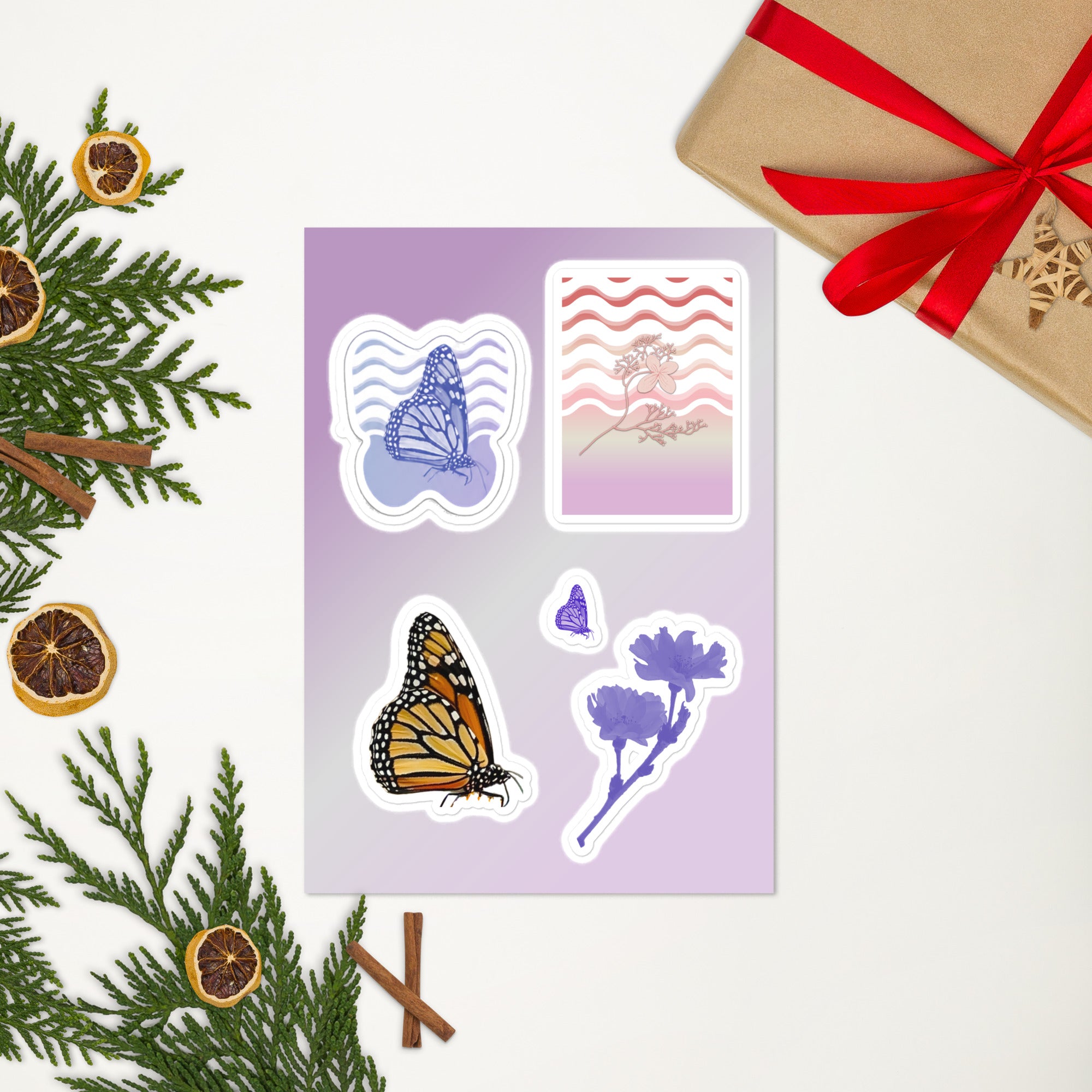 A sheet of decorative stickers displayed on a light background with holiday greenery, dried orange slices, cinnamon sticks, and a wrapped gift box. The stickers include a butterfly, a monarch butterfly, floral illustrations, and a patterned card design.