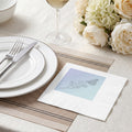 A white paper napkin with a printed butterfly and floral illustration, styled on a beige table runner at a dining table with white plates, a wine glass, and cream flowers.