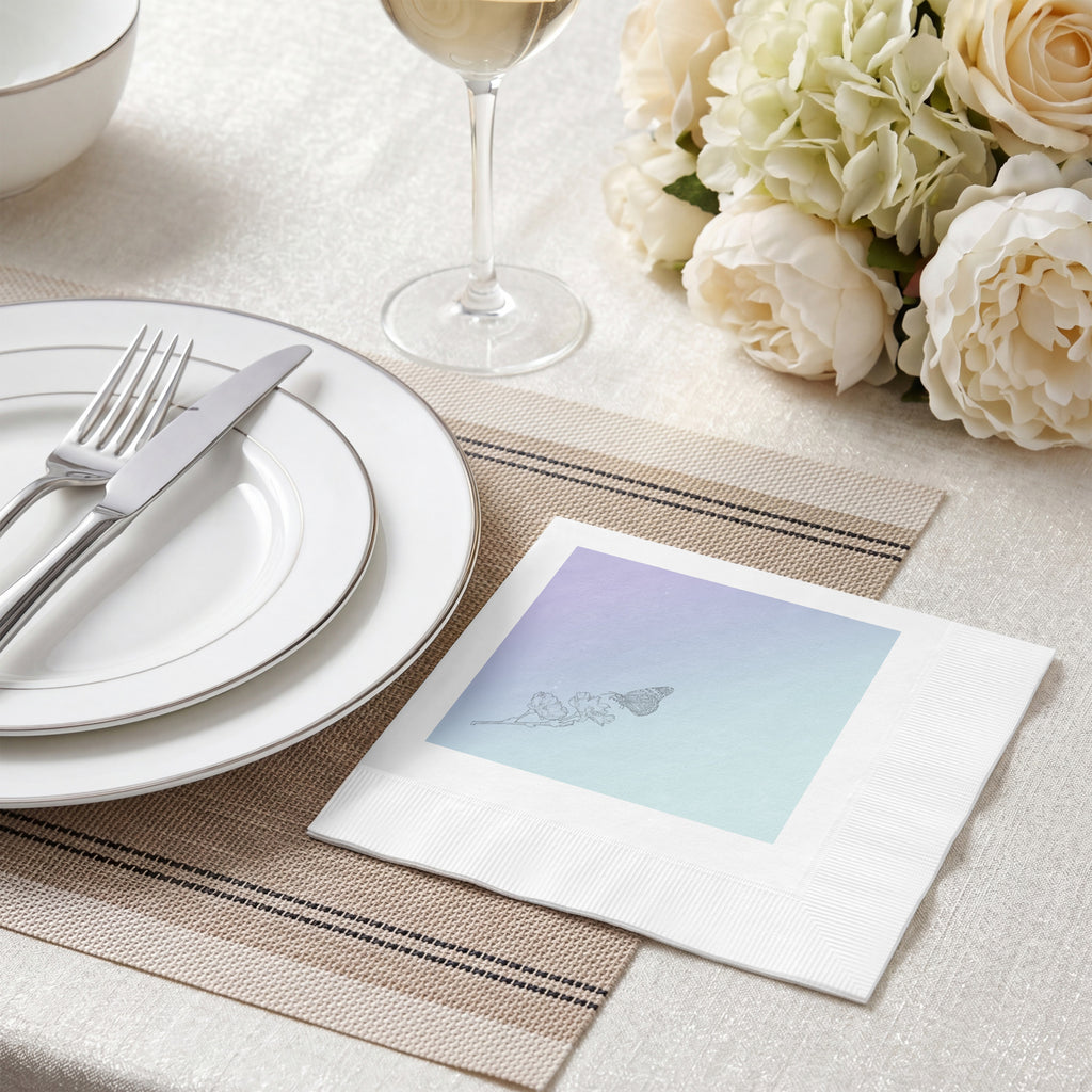 A white paper napkin with a pastel blue and purple gradient square print featuring a delicate line drawing of a butterfly, styled on a beige table setting with plates, a wine glass, and white flowers.