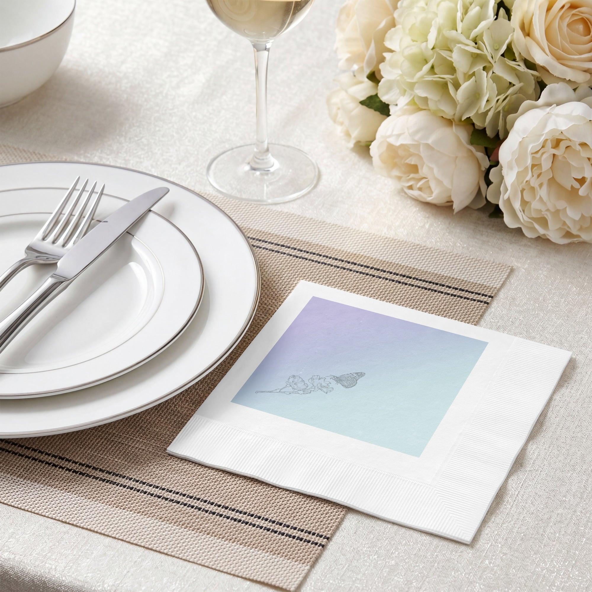 A white paper napkin with a pastel blue and purple gradient square print featuring a delicate line drawing of a butterfly, styled on a beige table setting with plates, a wine glass, and white flowers.